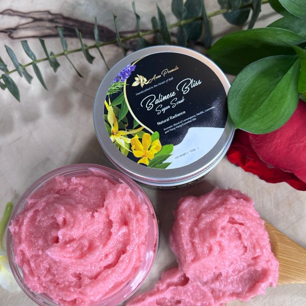 A container of Balinese Bliss Sugar Scrub with vibrant pink sugar crystals, scooped onto a wooden spatula. The jar label features a botanical-inspired design with lemongrass, vanilla, and ylang-ylang essential oils, promoting soft and radiant skin.