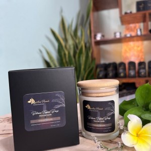 A glass jar of Balinese Tropical Breeze Massage Candle with a wooden lid, placed next to a sleek black gift box. The elegant label features tropical botanical illustrations, highlighting ylang-ylang, frangipani, sandalwood, and lemongrass essential oils. Soft lighting and delicate plumeria flowers enhance the spa-like ambiance.