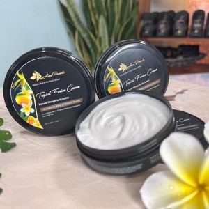 An open jar of Tropical Fusion Cream body butter with a silky, whipped texture, placed beside two closed jars featuring a mango and frangipani design on the label. The creamy formula is set against a tropical-themed background with natural flowers and wooden elements.