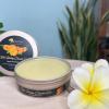 An open tin of Bali Healing Calendula Salve with a smooth, golden-yellow balm inside. The elegant packaging features bright calendula flowers on the label, emphasizing its soothing and skin-nourishing ingredients. The tin is placed on a wooden surface with tropical plumeria flowers beside it.