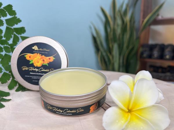 An open tin of Bali Healing Calendula Salve with a smooth, golden-yellow balm inside. The elegant packaging features bright calendula flowers on the label, emphasizing its soothing and skin-nourishing ingredients. The tin is placed on a wooden surface with tropical plumeria flowers beside it.